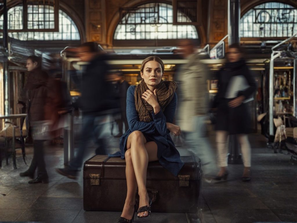 Woman sits on a vintage brown suitcase in a busy train station, surrounded by blurred pedestrians rushing by.