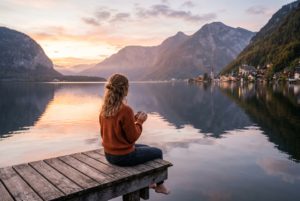 Woman in an orange sweater sits on a weathered wooden dock, holding a mug, overlooking a calm lake at sunset with mountains and a village in the distance.