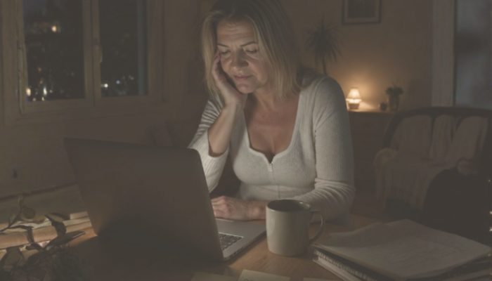 Woman working late at night at a laptop, resting her chin on her hand, with a mug and notes on the desk in a dimly lit room.